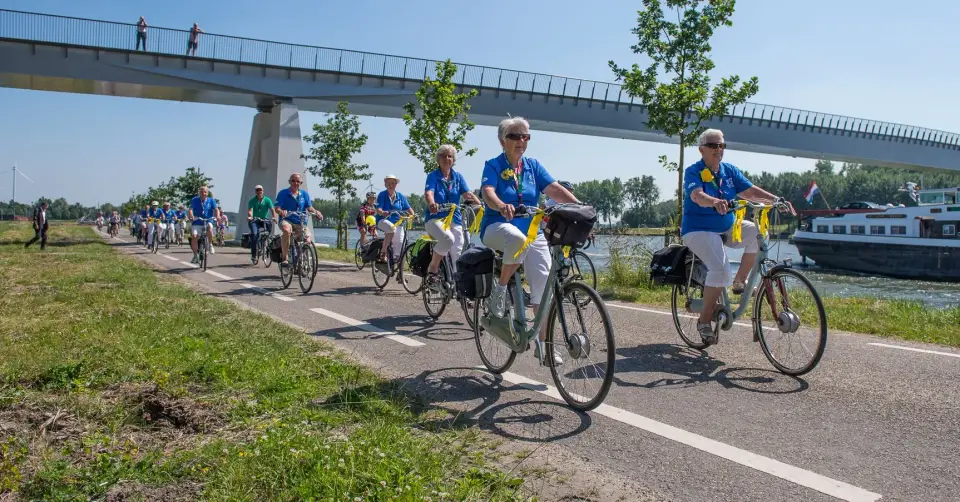 Een lange rij recreanten in blauwe poloshirts met gele linten fietst over een fietspad langs het kanaal, onder een fietsbrug door