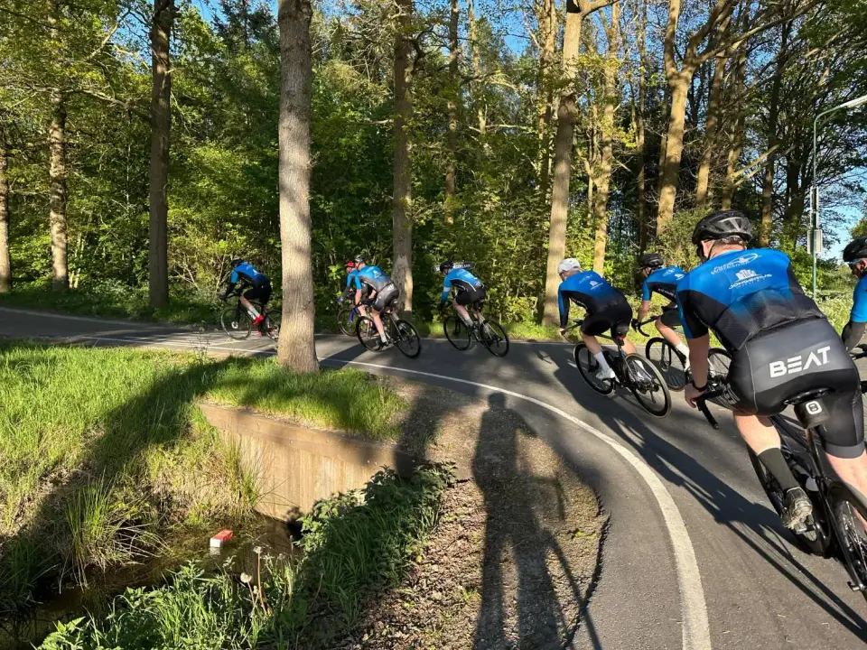 Toerleden in het blauwe clubtenue fietsen achter elkaar door een bocht langs de bosrand, in de avondzon