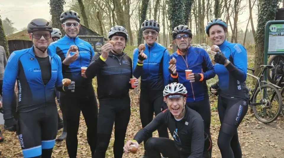 Lachende toerleden in winterkleding en met helm houden tijdens een gravelrit in het bos een oliebol omhoog